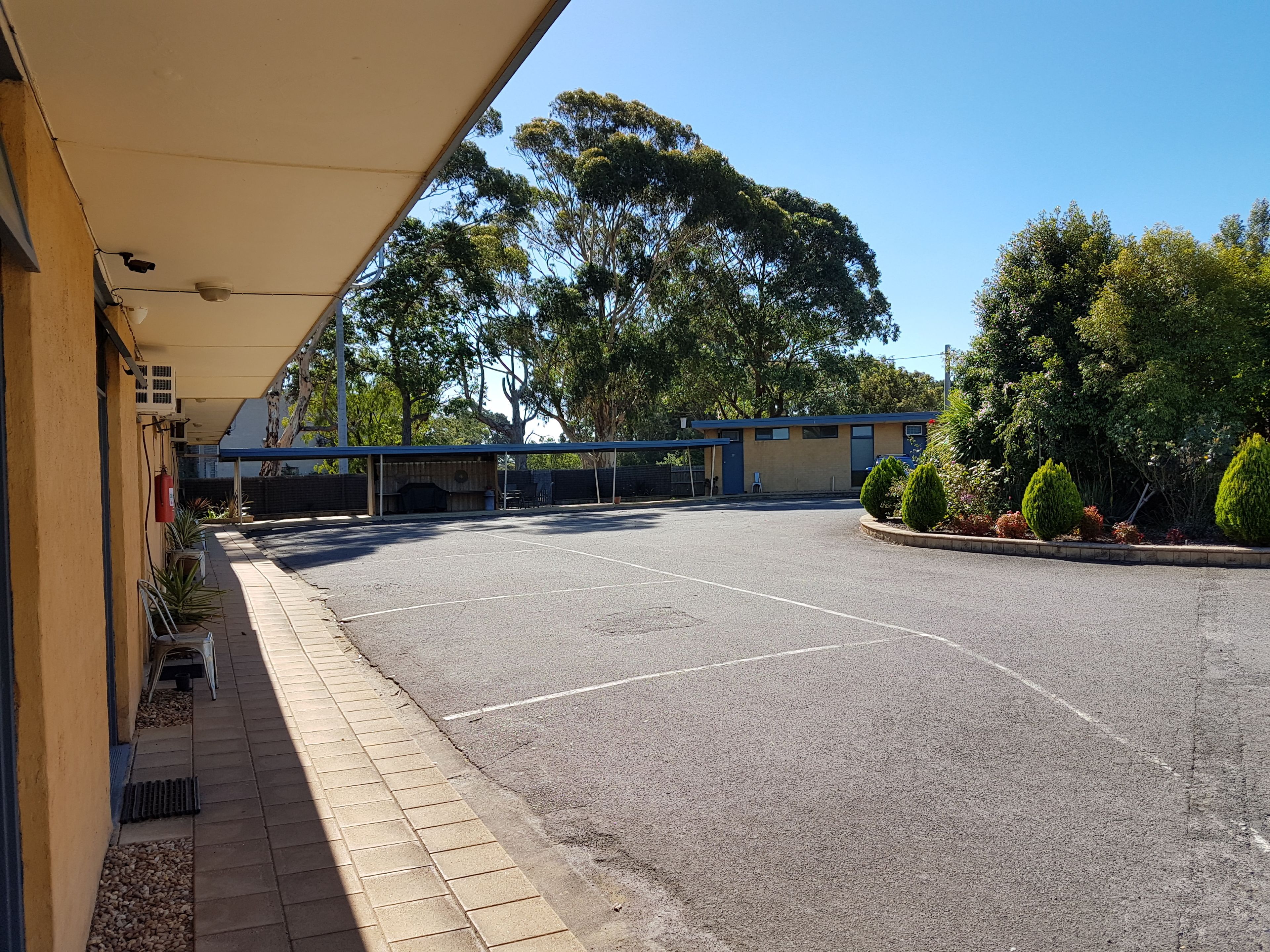 A parking lot with a building on the left, covered by a roof, and a row of potted plants.