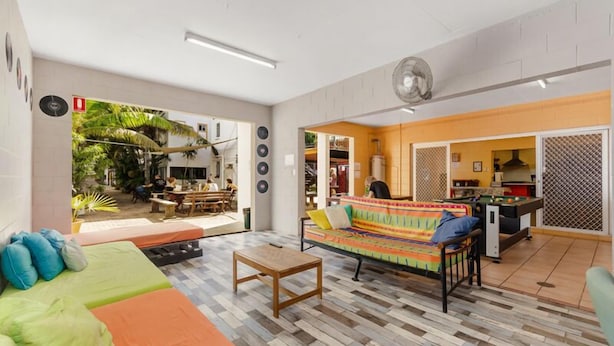 Amplia sala de estar con sofá, mesa de centro y un sofá colorido. Se observa un área de cocina con mesa y sillas, y vista a un jardín con palmeras.