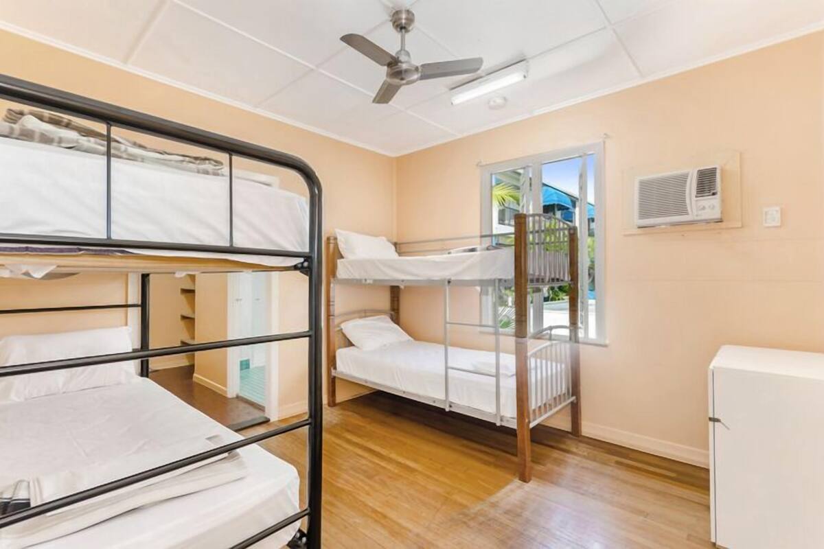 Une chambre de dortoir avec des lits superposés, un ventilateur de plafond, une fenêtre donnant sur l’extérieur et un réfrigérateur blanc.