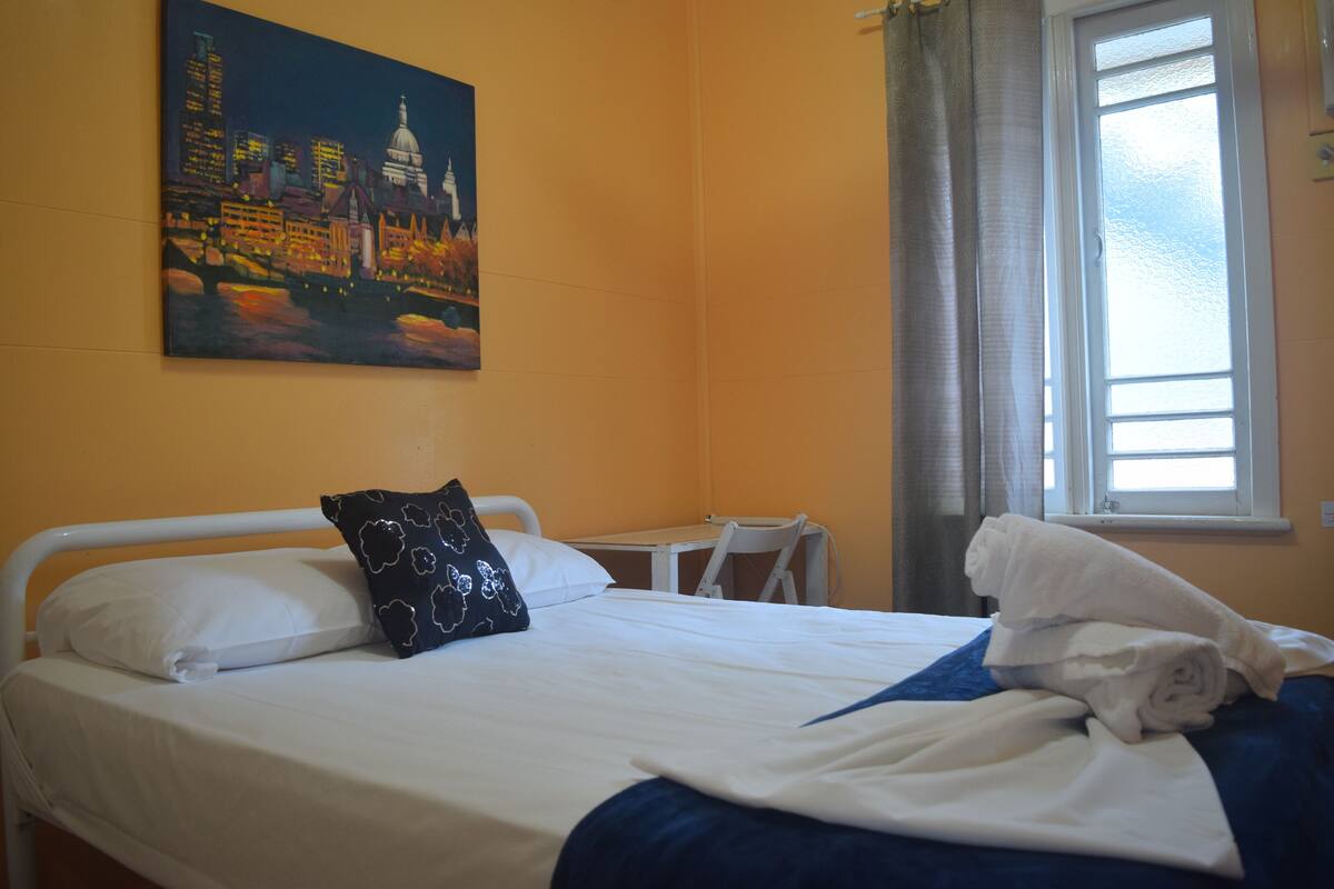 Une chambre d’hôtel avec un lit, un tableau représentant un paysage urbain, une fenêtre avec des rideaux et une petite table.