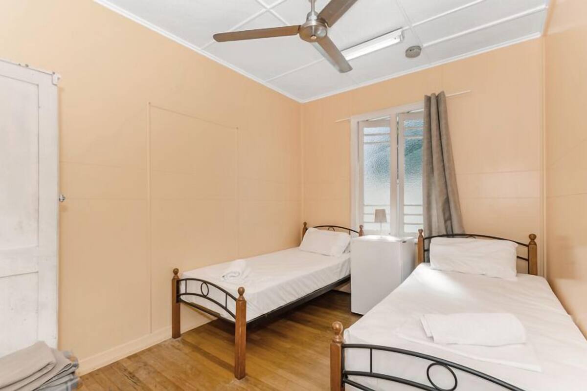 Une chambre avec deux lits simples, un ventilateur de plafond, une fenêtre avec des rideaux et une porte.