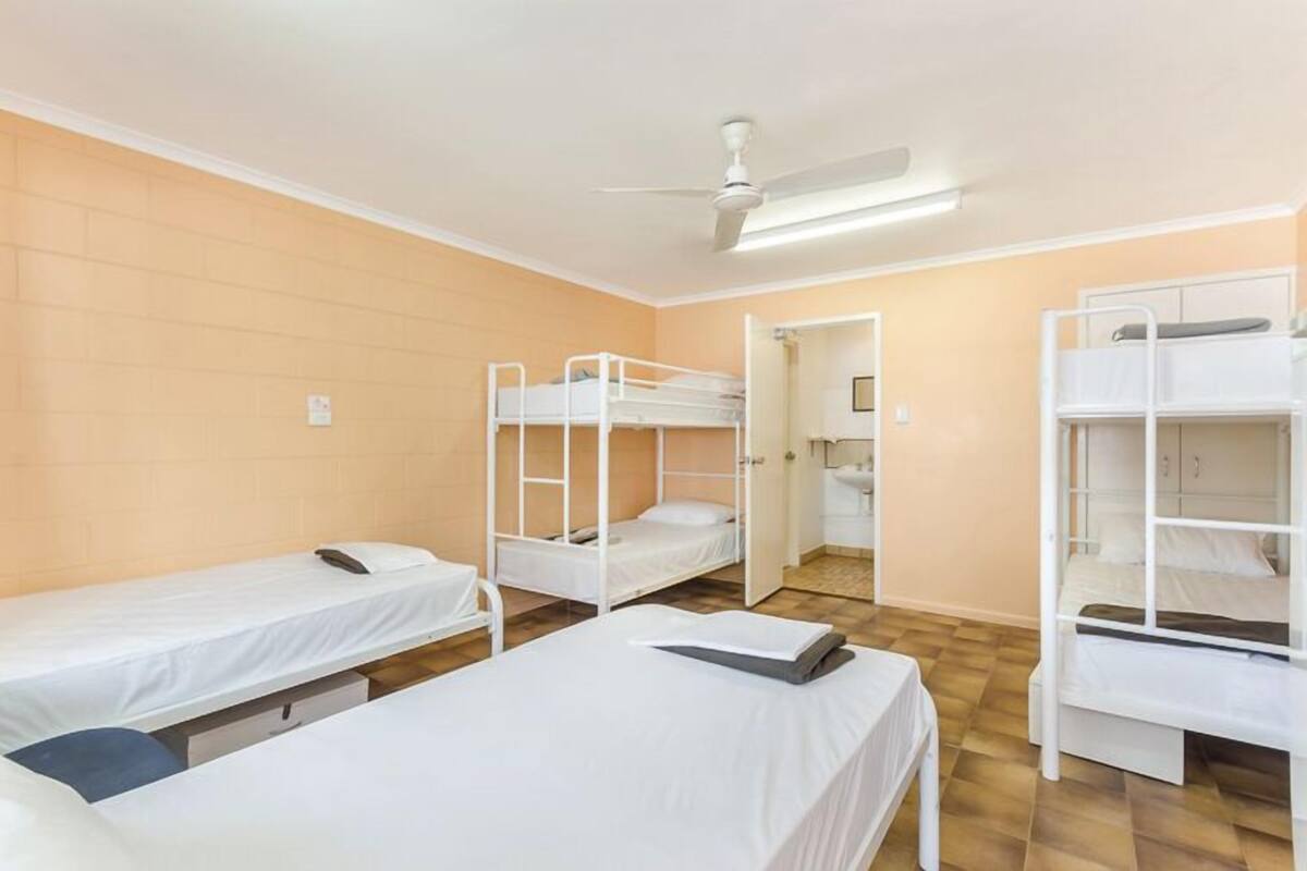 Une chambre de dortoir avec des lits superposés, un ventilateur de plafond et une petite salle de bain visible à travers une porte ouverte.