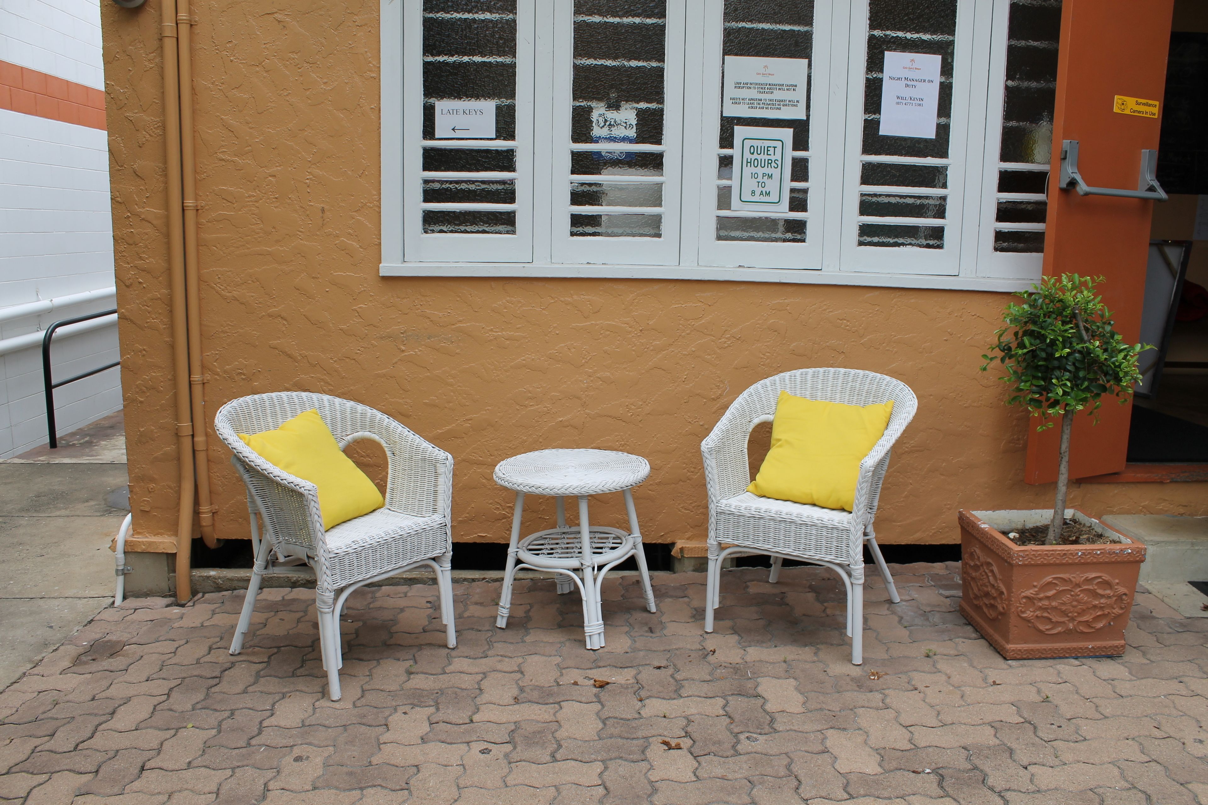 A small outdoor seating area with two white wicker chairs and a round table, each with a yellow cushion. There is a potted plant to the right.