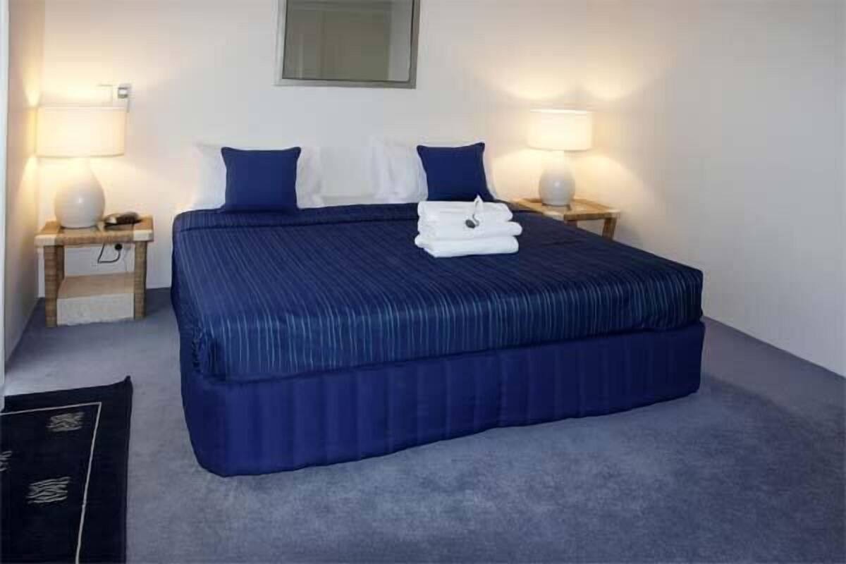 Ein ordentlich bezogenes Bett mit blauer Bettwäsche und weißen Kissen, zwei Nachttischlampen und ein gefaltetes Handtuch auf dem Bett.