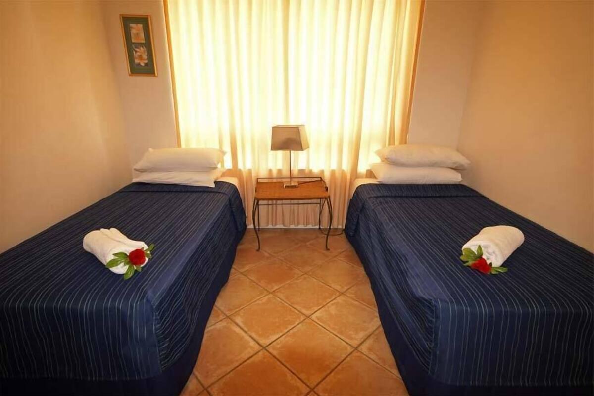 Ein Hotelzimmer mit zwei Betten, jedes mit einem Kopfkissen und einem Handtuch, sowie einem kleinen Tisch mit einer Lampe dazwischen.