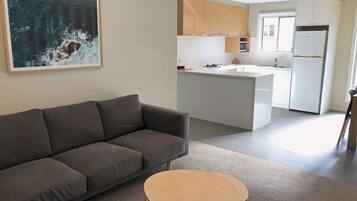 Ein modernes Wohnzimmer mit einem grauen Sofa und einem kleinen runden Tisch, eine Küche mit weißen Schränken und Geräten, sowie ein großes abstraktes Gemälde an der Wand.