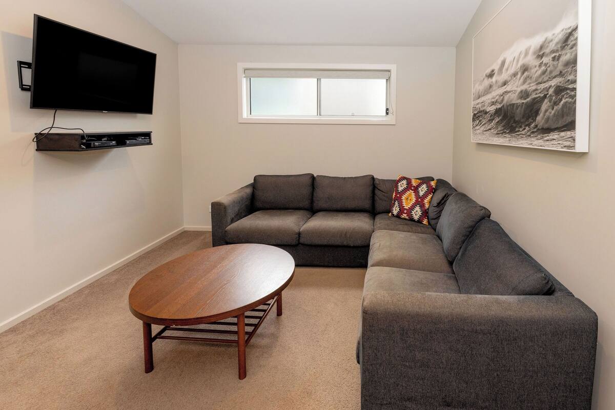 Een woonkamer met een hoekbank, een flatscreen televisie aan de muur, een raam en een ingelijst kunstwerk.