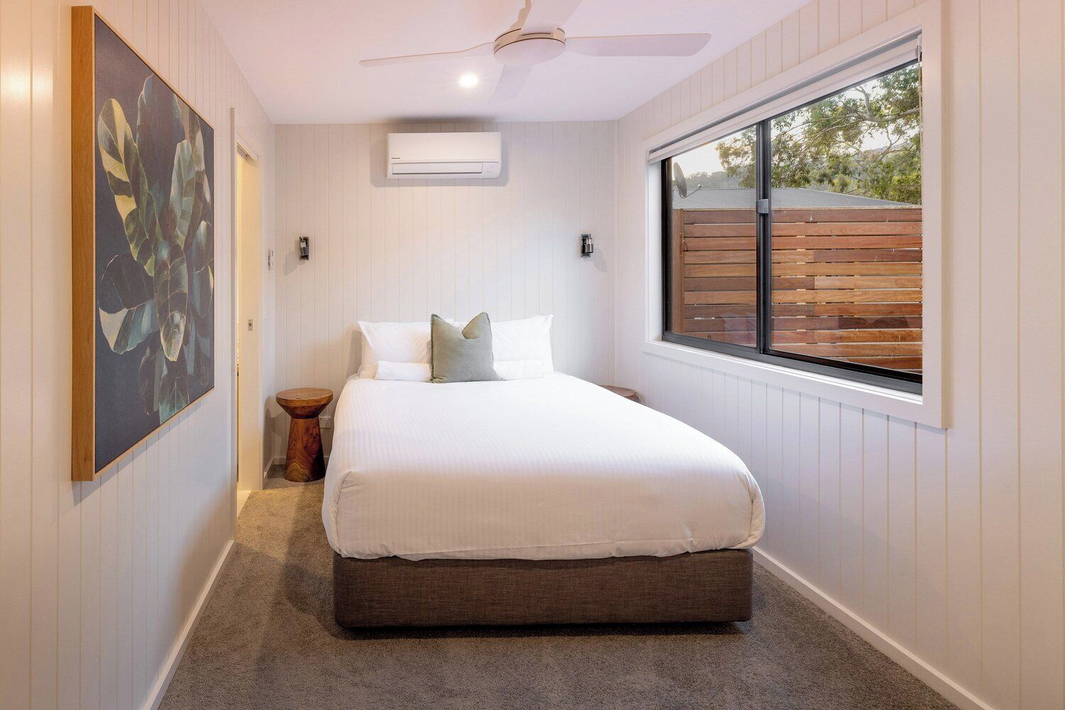 A bedroom with a bed, a ceiling fan, an air conditioning unit, a window with blinds, and a wooden floor.