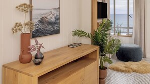 Ein Zimmer mit einem Holzkonsolentisch, einem großen gerahmten Bild des Meeres, einem Fernseher und Blick auf das Meer durch ein Fenster.