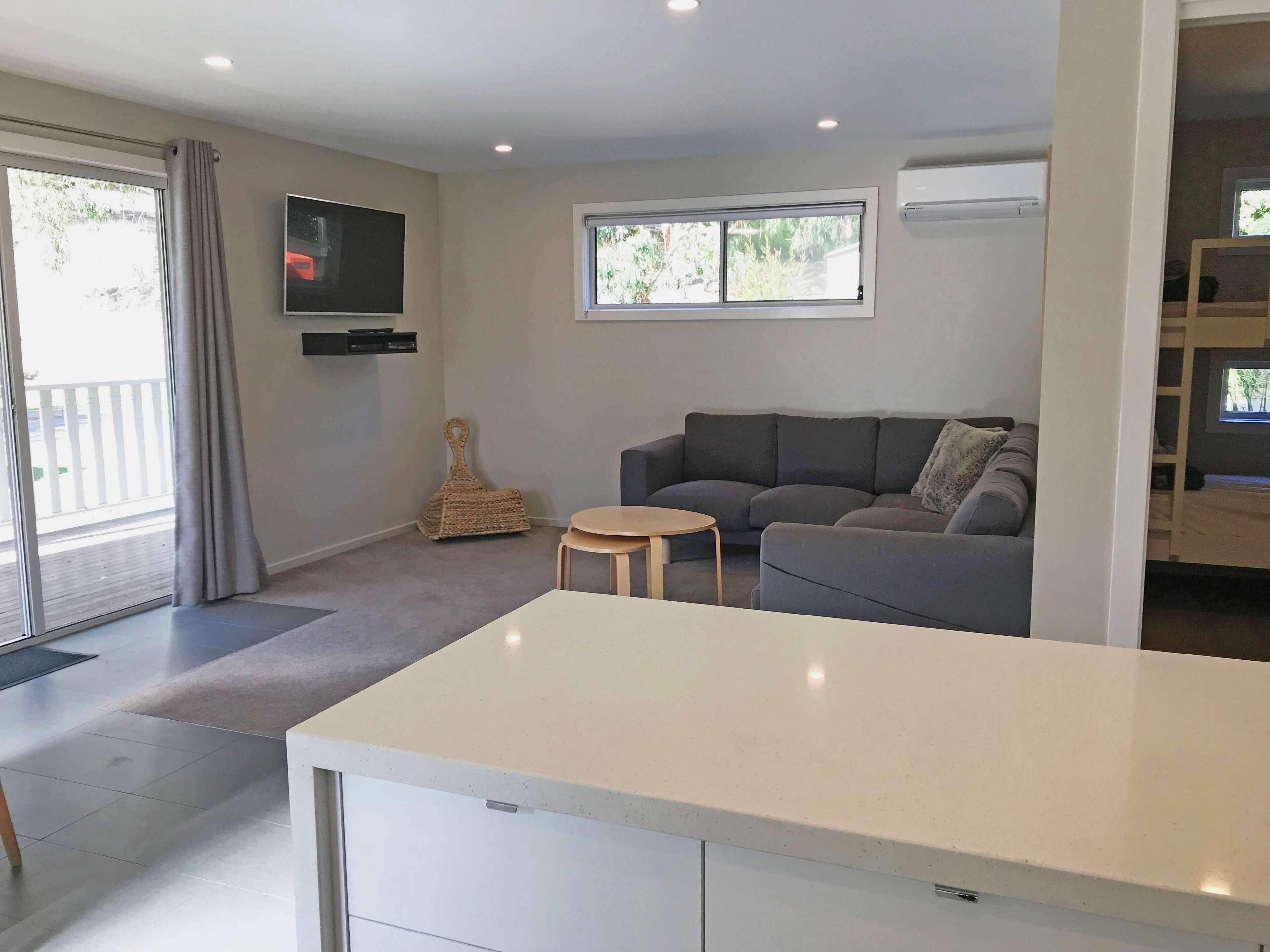 Una sala moderna con un sofá gris, una mesita redonda, un televisor montado en la pared y una ventana con vista a la vegetación.