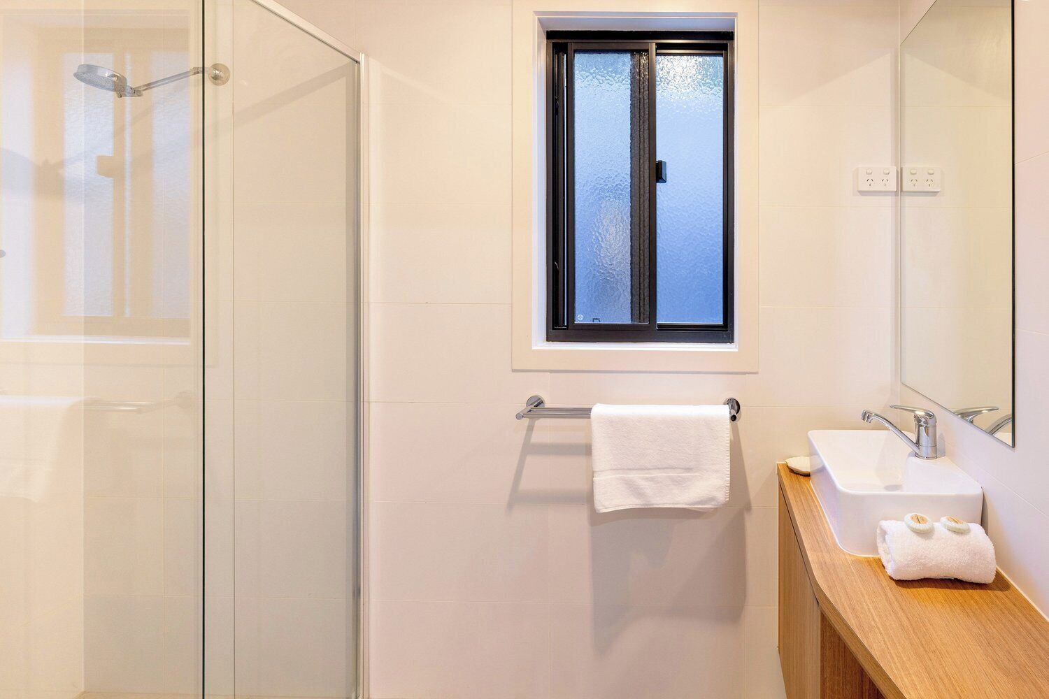 Ein modernes Badezimmer mit Duschkabine aus Glas, einem Fenster über dem Waschbecken und einem Holzwaschtisch.