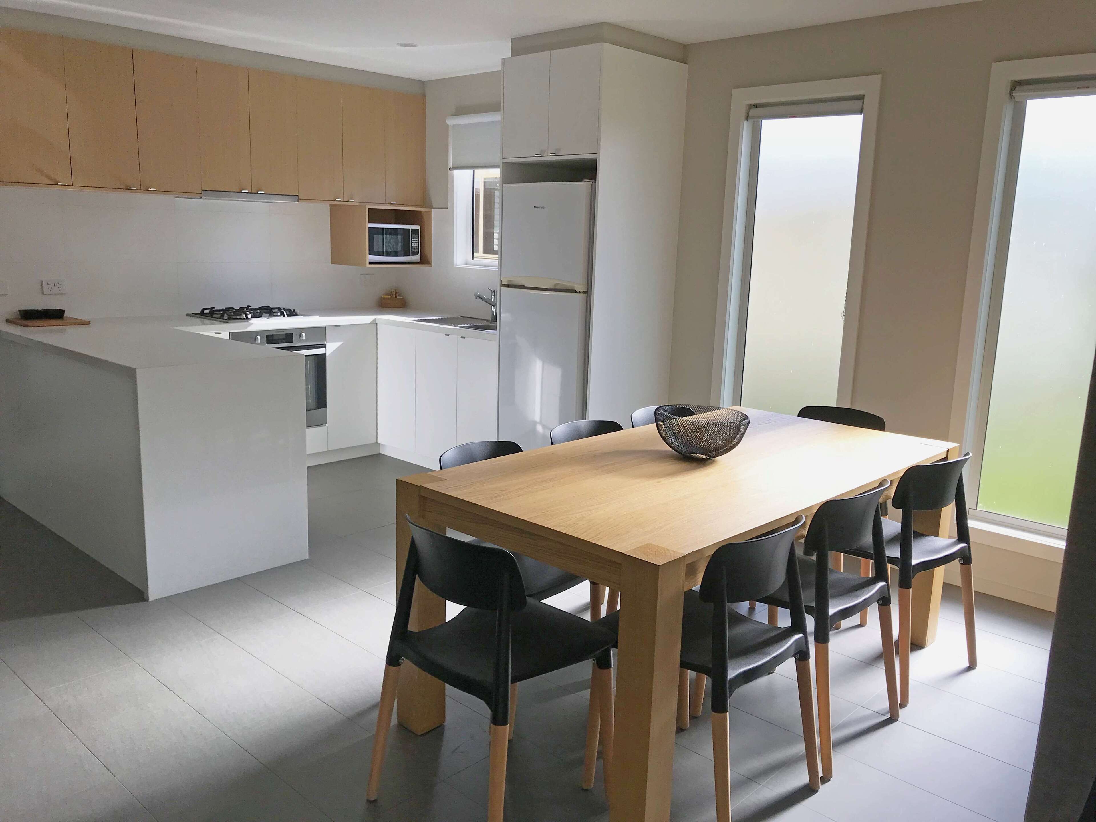 A modern kitchen with a central island, a dining table with chairs, and a large window.