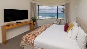 Ein Schlafzimmer mit einem großen Bett, einem Fernseher auf einer Holzkonsole, Blick aufs Meer und einer Pflanze.