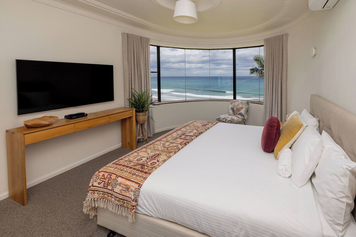 Een slaapkamer met een groot bed, een tv op een houten dressoir, uitzicht op de oceaan en een plant.