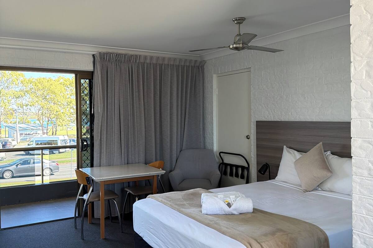 A hotel room with a bed, a desk, a chair, a ceiling fan, and a view of the outside.