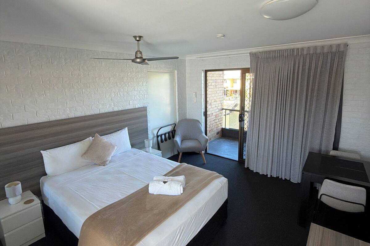 A hotel room with a bed, a desk, a chair, a ceiling fan, and a view of the outside.