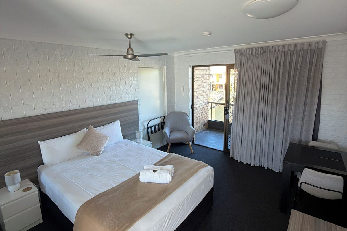 A hotel room with a bed, a desk, a chair, a ceiling fan, and a view of the outside.