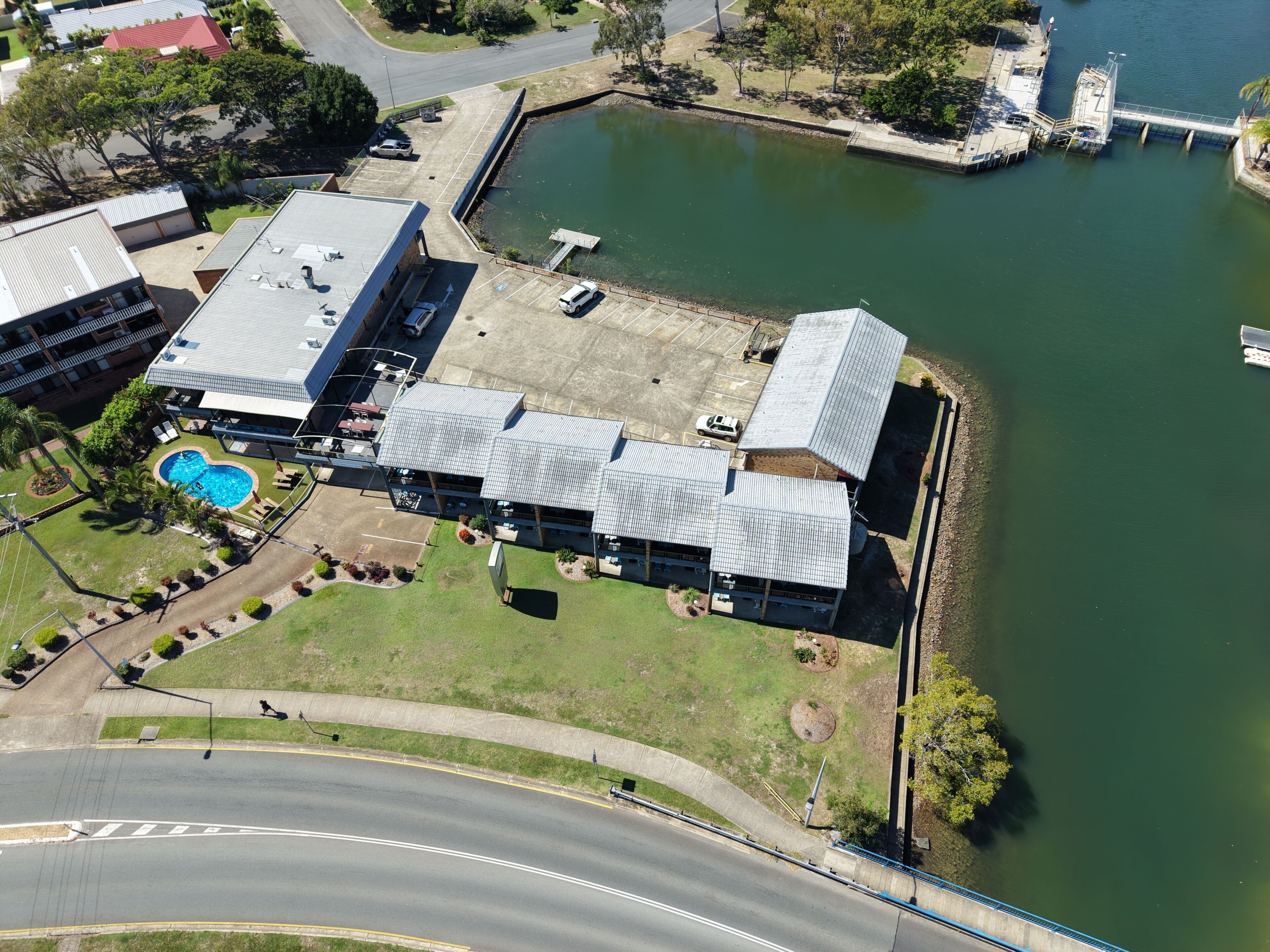An aerial view of a building with a swimming pool, parking lot, and a dock extending into a body of water.