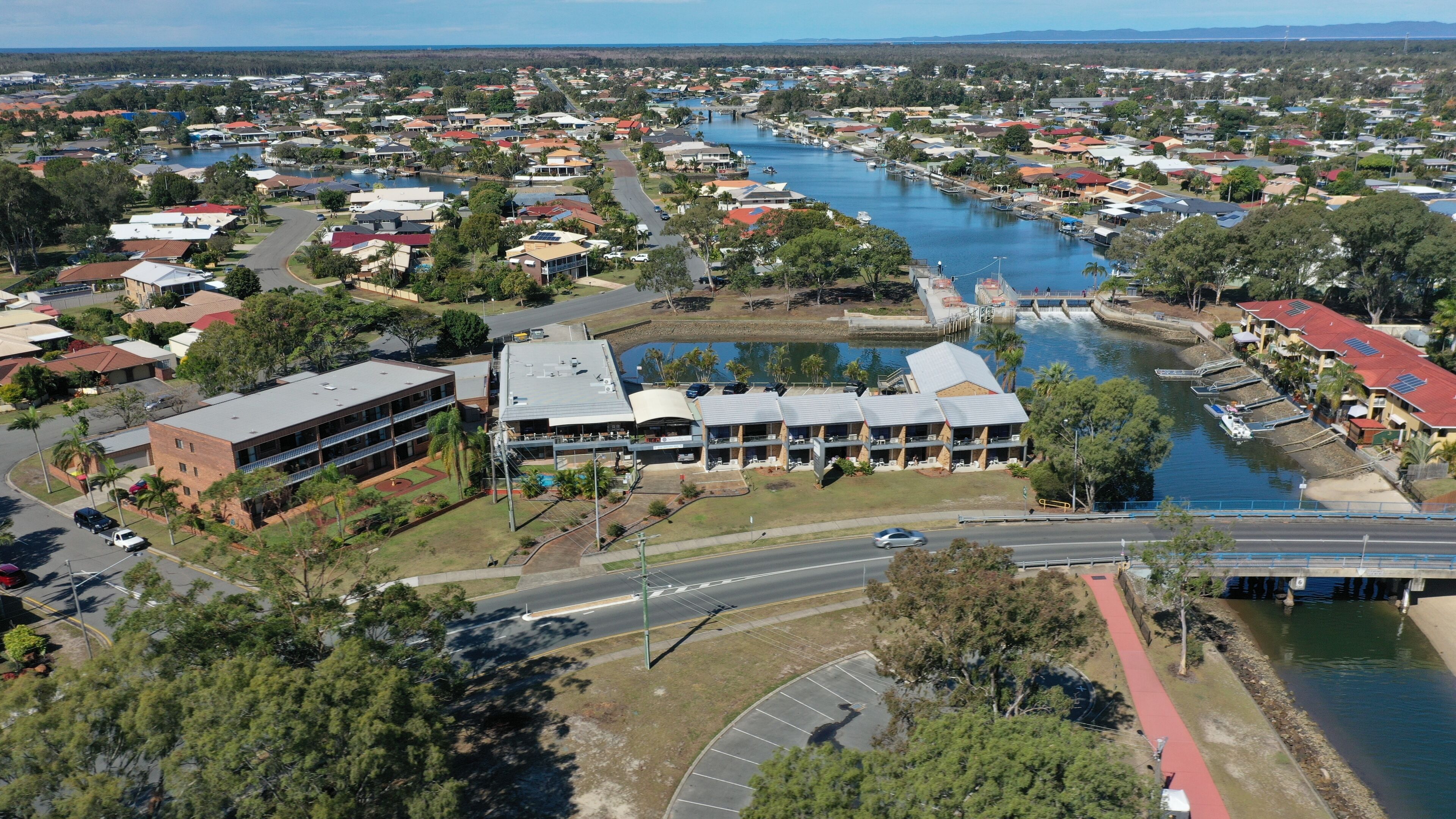 An aerial view of a coastal town with a marina, residential buildings, and a prominent hotel with a large pool and multiple floors.