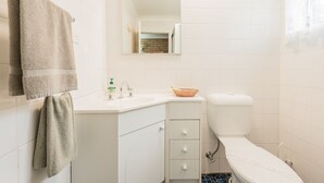 A bathroom with white tiles, a sink with a basket, a toilet, and a mirror.