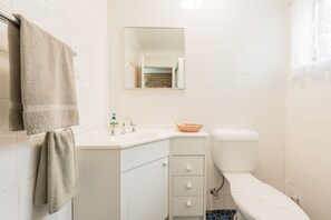 A bathroom with white tiles, a sink with a basket, a toilet, and a mirror.