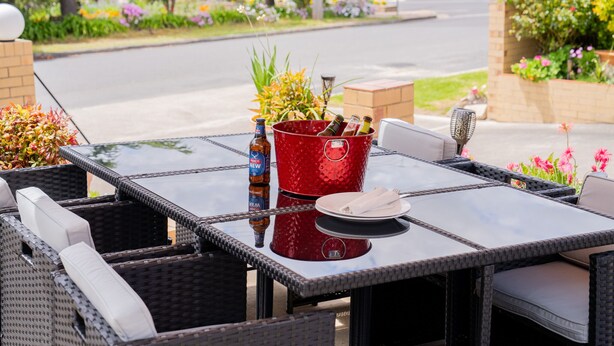 An outdoor patio with a glass table, wicker chairs, and a red bucket with bottles. - Settlers Motor Inn (Tenterfield)