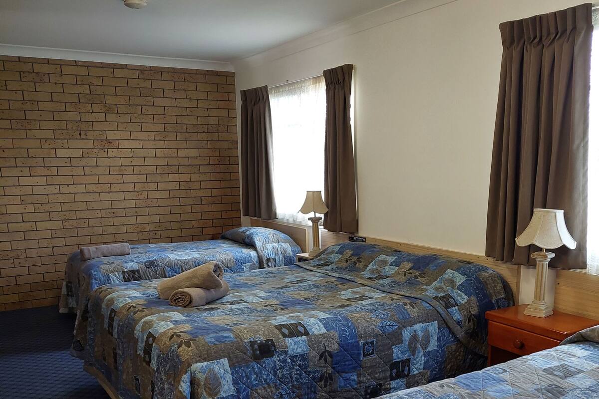 Habitación de hotel con dos camas, una pared de ladrillo, una ventana con cortinas y una mesita de noche con lámpara.