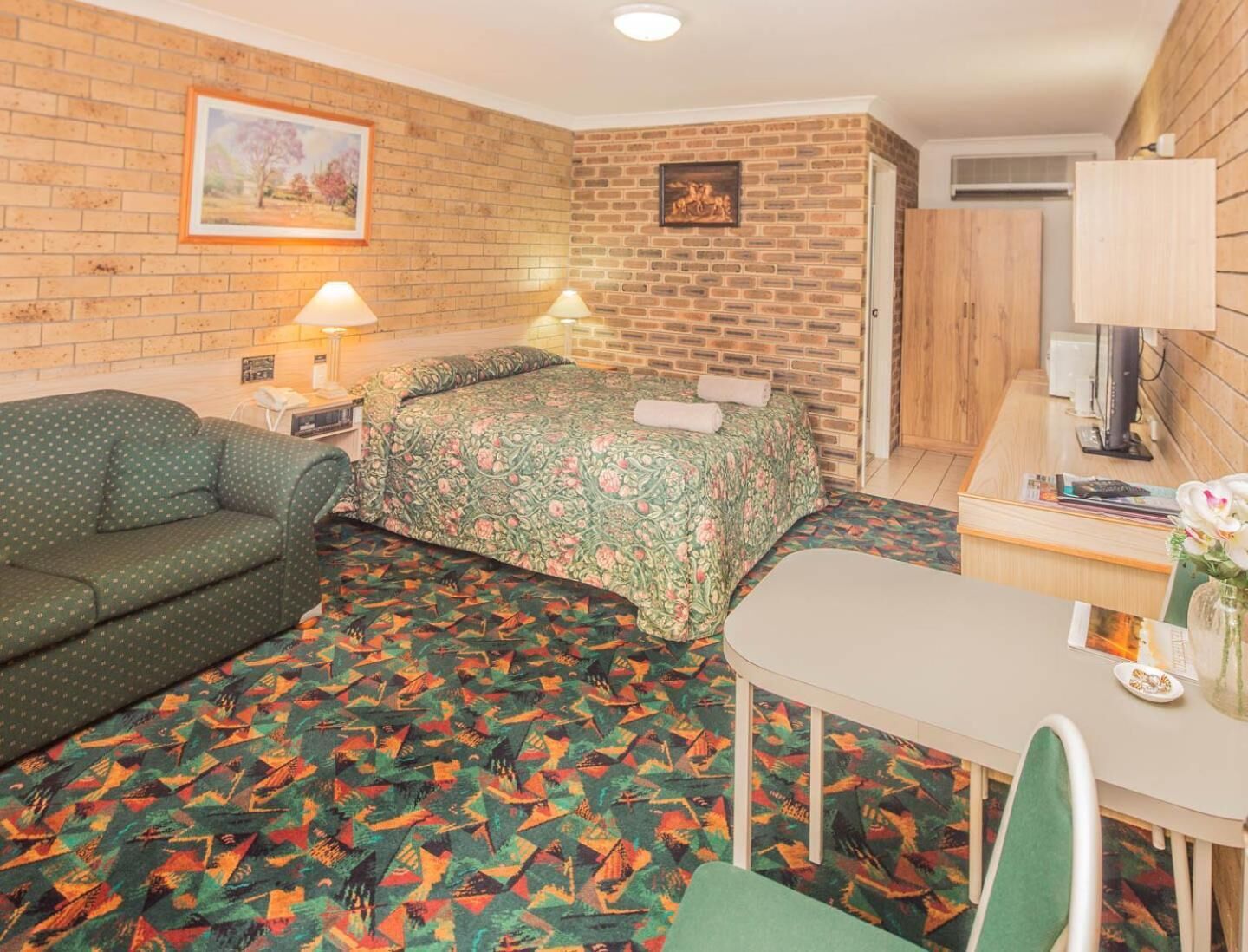 A room with a brick wall, a bed with floral bedding, a green sofa, a small table with a lamp, and a television.