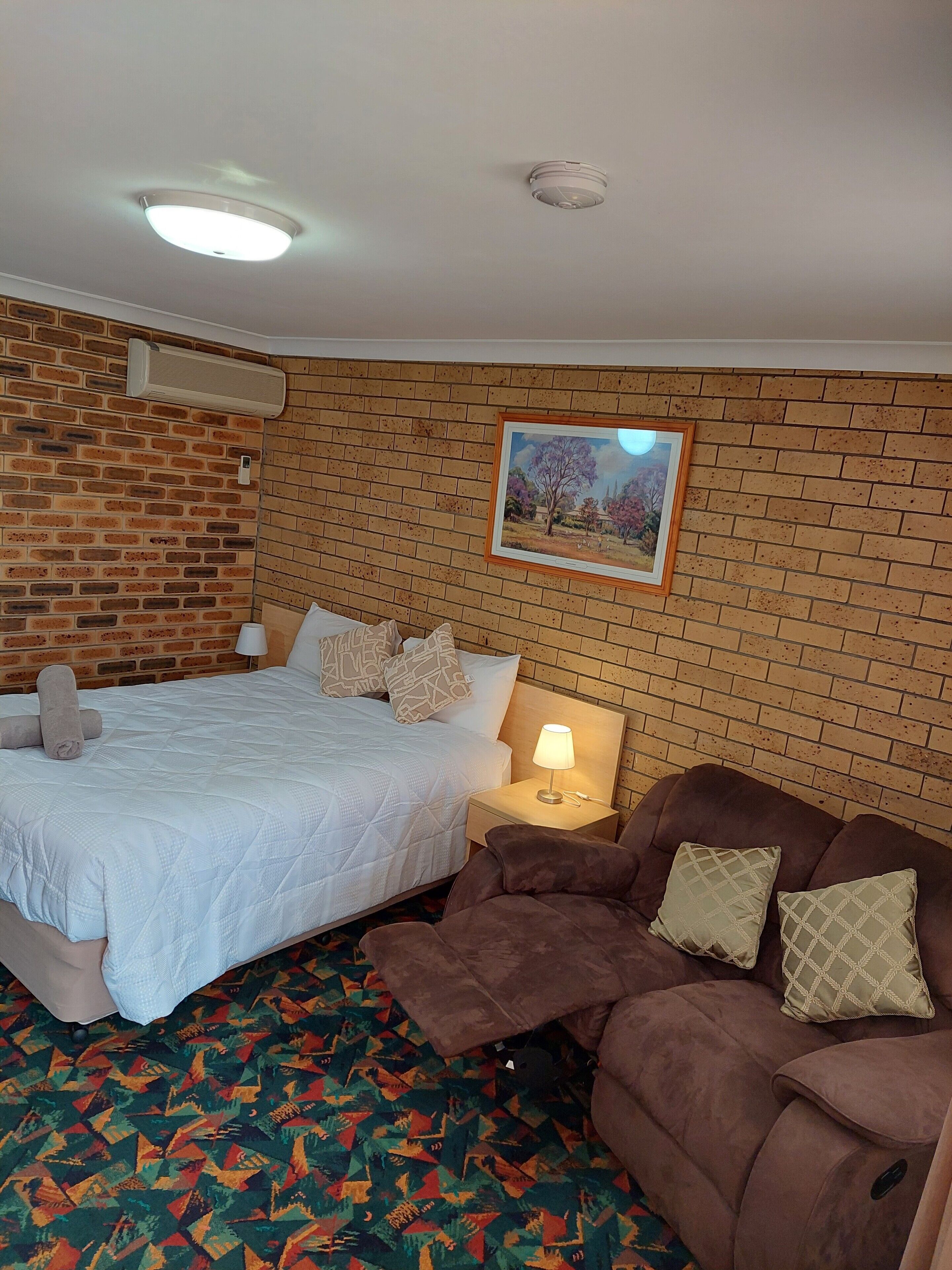 A hotel room with a brick wall, a bed with white bedding, a brown sofa with patterned cushions, a nightstand with a lamp, and a framed picture on the wall.