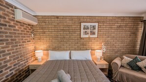 A hotel room with a brick wall, a large bed with a quilted bedspread, two bedside lamps, a framed picture on the wall, and a sofa with cushions. - Settlers Motor Inn (Tenterfield)