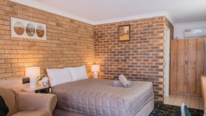 A hotel room with a brick-patterned wall, a bed with a grey quilt, a wooden wardrobe, and a small table with a lamp and a clock.