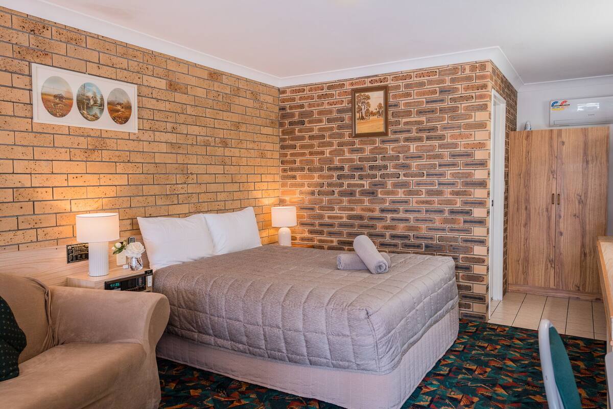 Una habitación de hotel con pared de ladrillo, cama con colcha gris, armario de madera y pequeña mesa con lámpara y reloj.