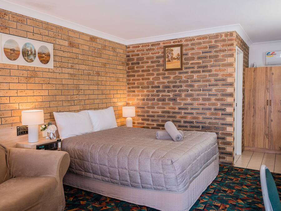Una habitación de hotel con pared de ladrillo, cama con colcha gris, armario de madera y pequeña mesa con lámpara y reloj.