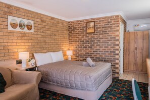A hotel room with a brick-patterned wall, a bed with a grey quilt, a wooden wardrobe, and a small table with a lamp and a clock.