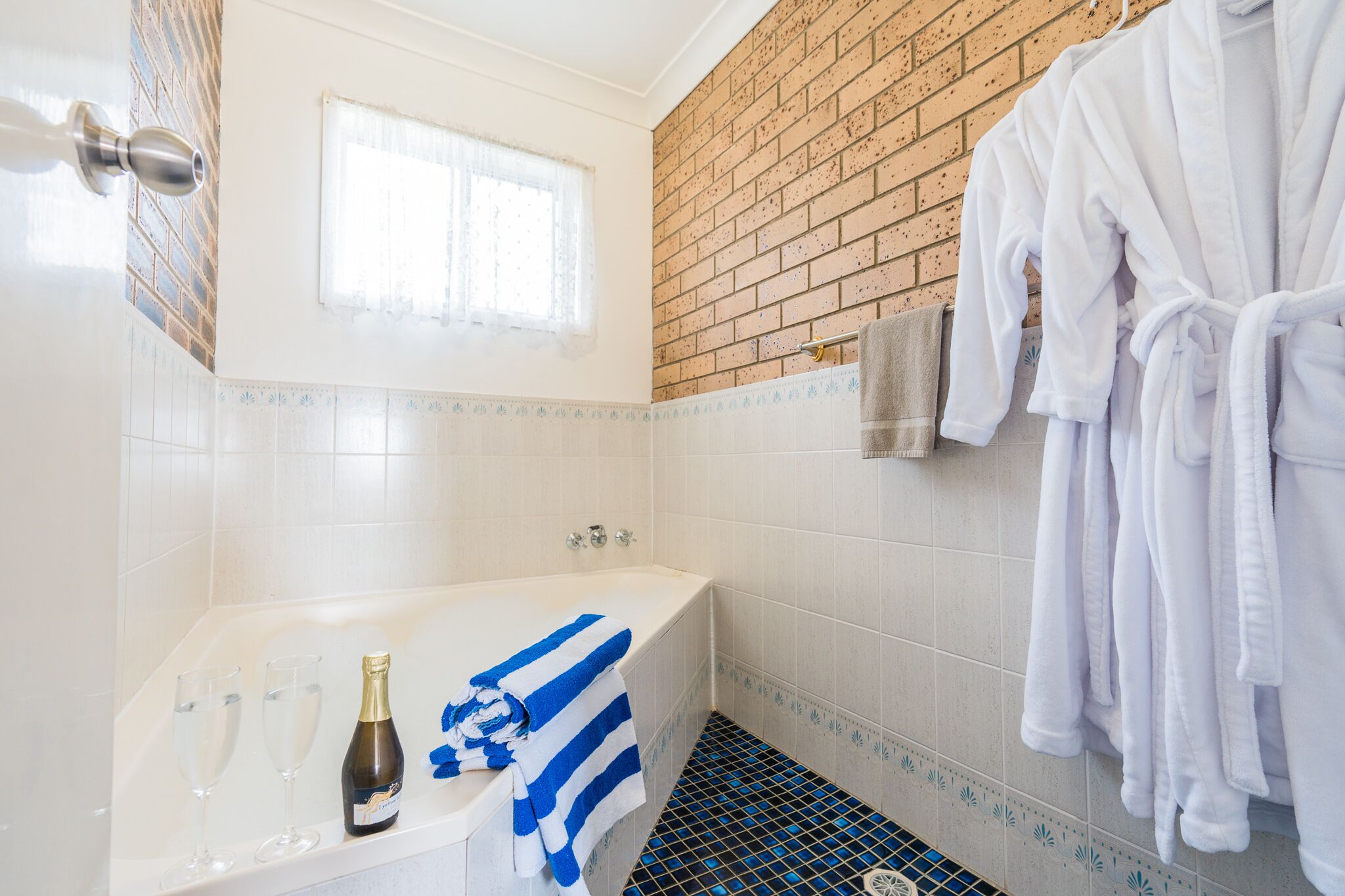 A bathroom with a brick wall, a white bathtub, a towel rack with a towel, and a bottle of champagne.