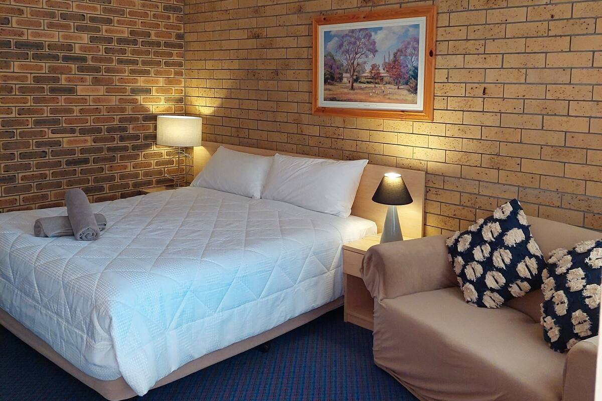 Habitación de hotel con pared de ladrillo, una cama con colcha, mesita de noche con lámpara, sofá con cojines estampados y un cuadro en la pared.