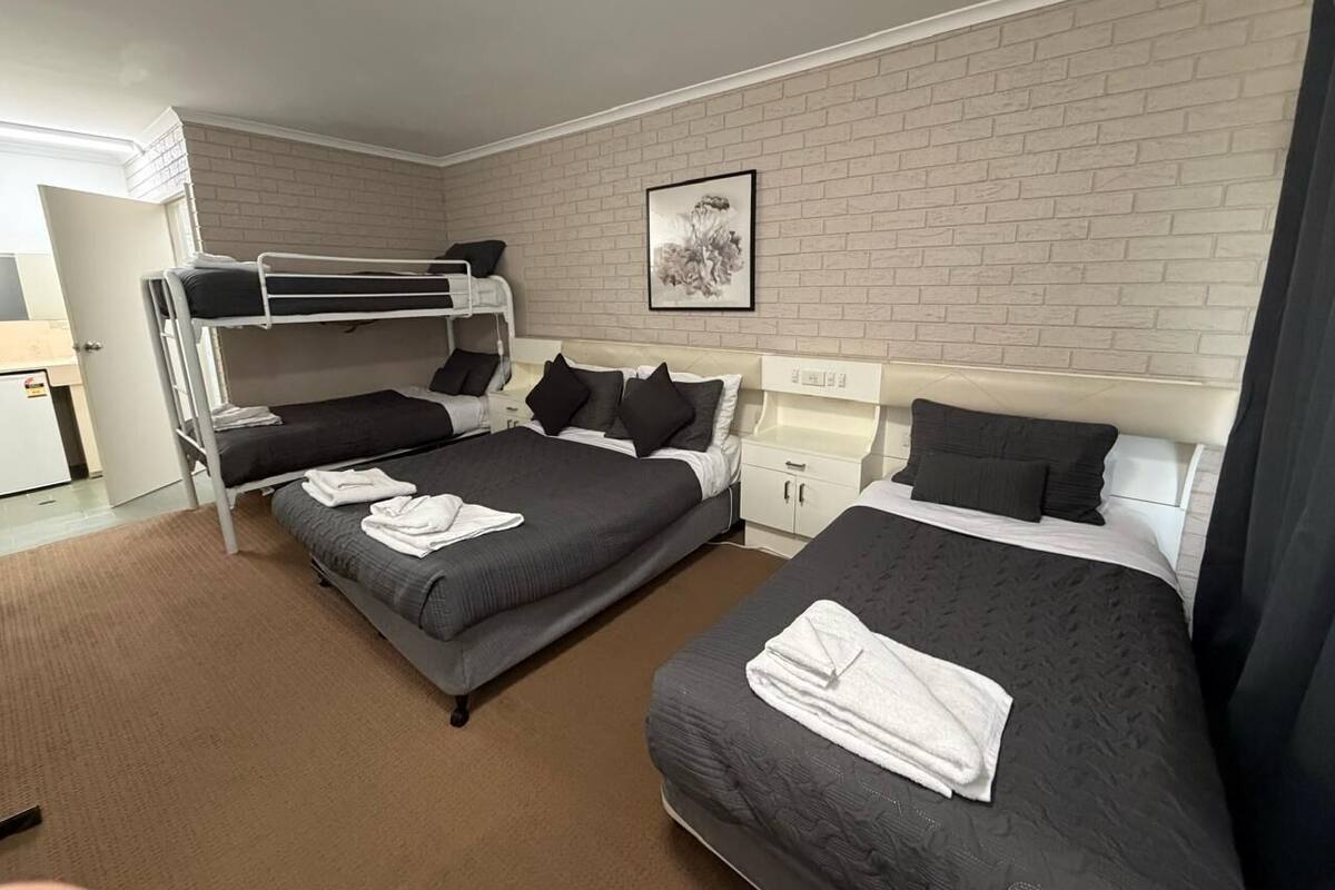 Ein Hotelzimmer mit zwei Betten, einem Etagenbett, einem kleinen Schreibtisch und einer weißen Ziegelwand.
