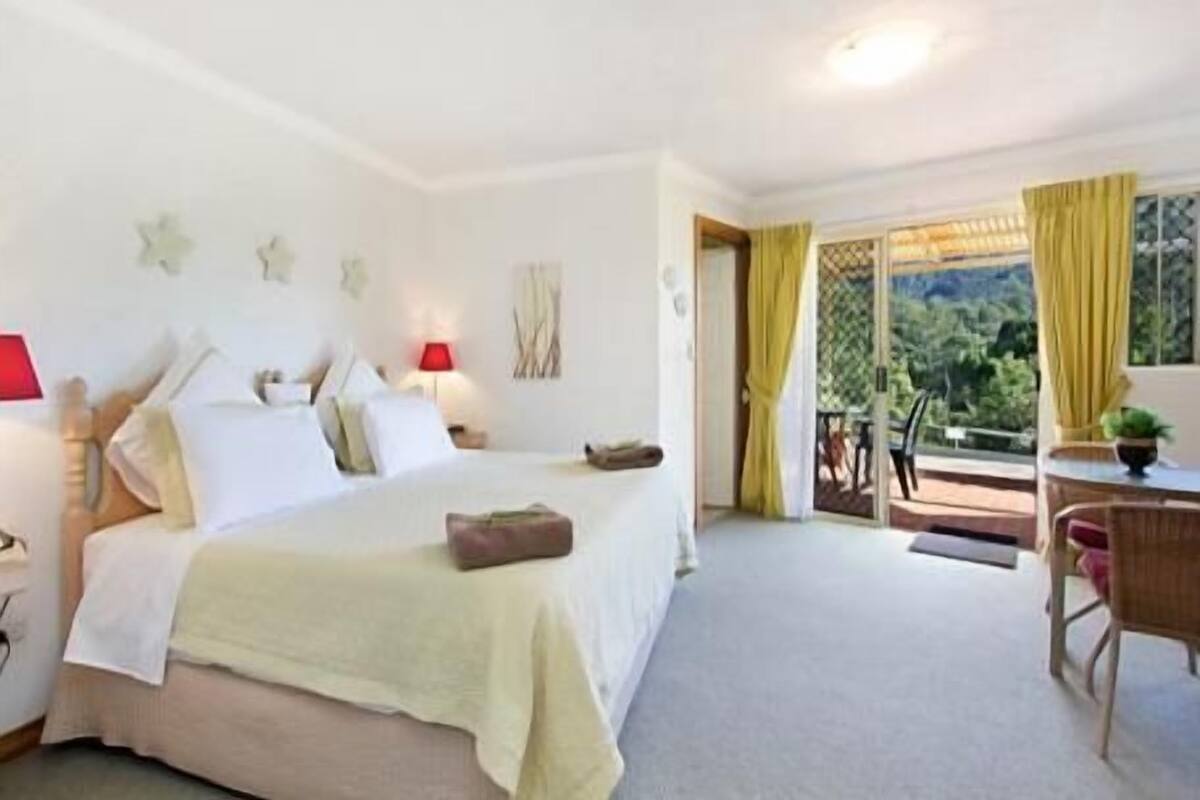 A bedroom with a bed, bedside tables, a desk, and a chair. There is a window with curtains and a view of greenery outside.