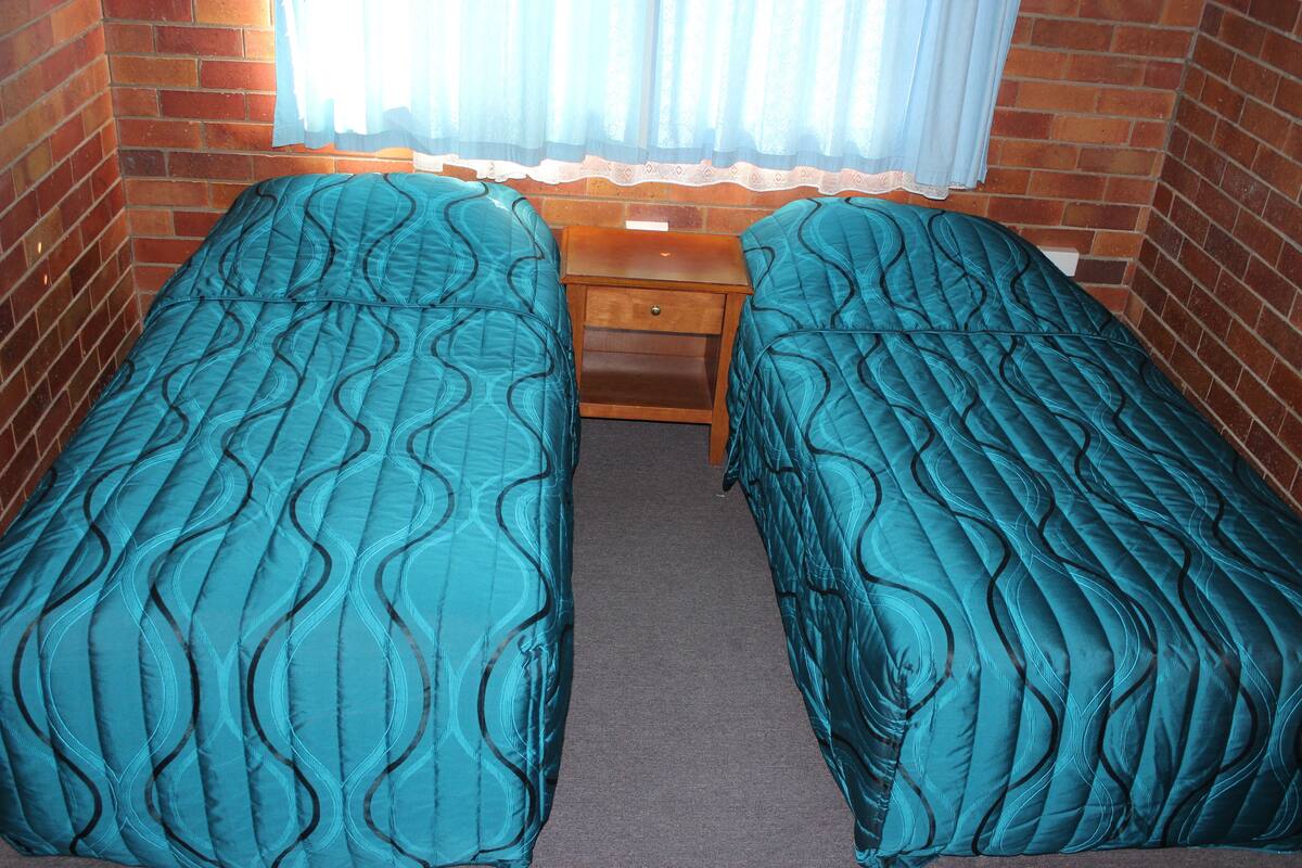 A room with two single beds, a wooden nightstand, and a window with sheer curtains.