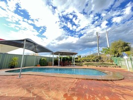 Une piscine avec un patio en pierre, un espace salon couvert et une clôture verte.