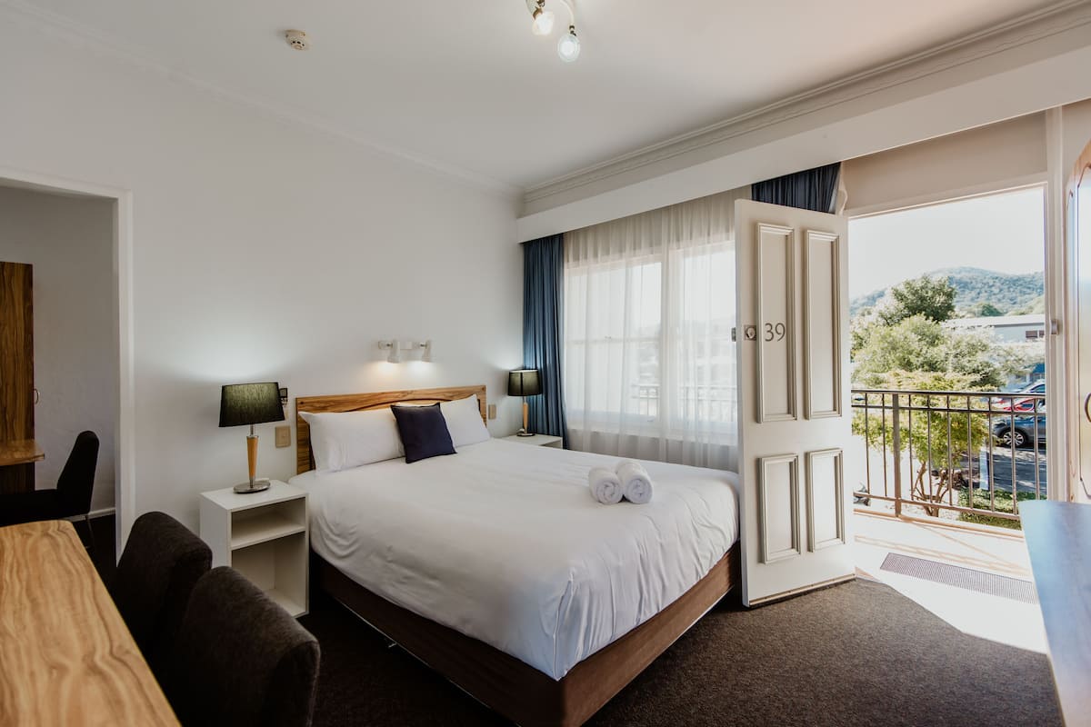 A hotel room with a large bed, a desk, a chair, a nightstand with a lamp, and a view of the outside.