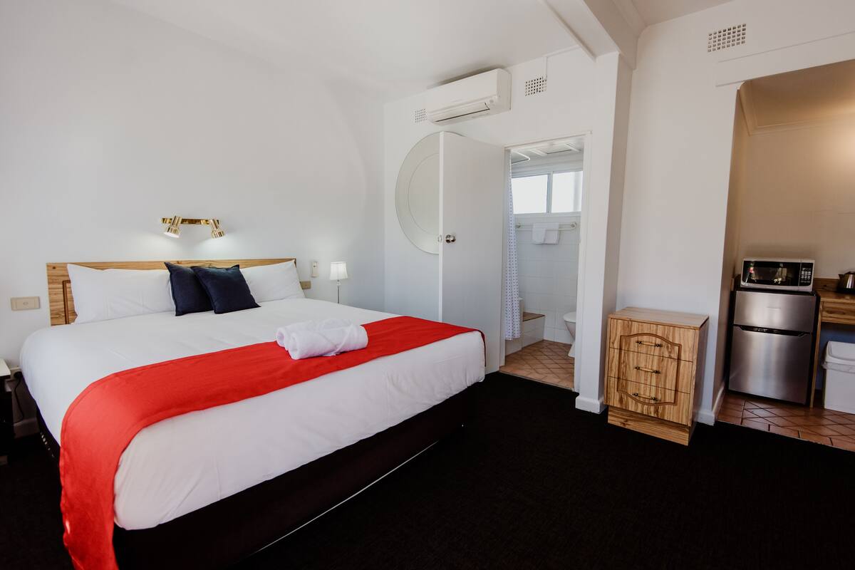 A hotel room with a large bed, a small kitchenette, an air conditioning unit, and a bathroom visible through an open door.