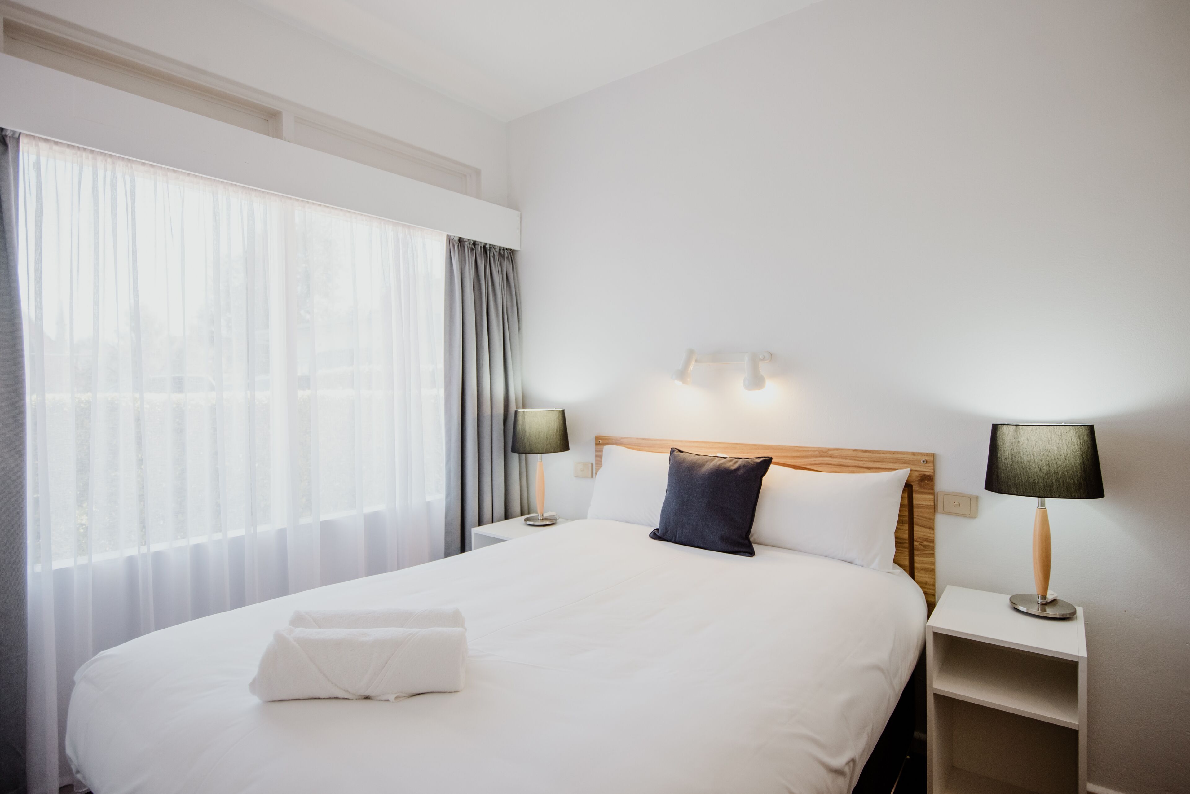 A hotel room with a large bed, two bedside tables with lamps, a wooden headboard, and sheer curtains.