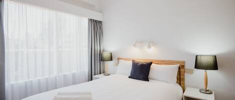 A hotel room with a large bed, two bedside tables with lamps, a wooden headboard, and sheer curtains.