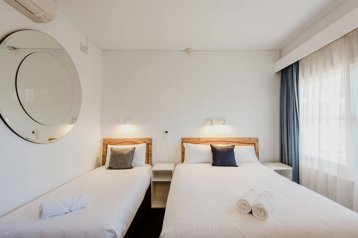 A hotel room with two beds, a round mirror, bedside lamps, and a window with curtains.