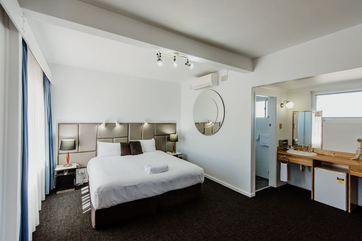 A hotel room with a large bed, a round mirror, a wooden vanity, and a small refrigerator.