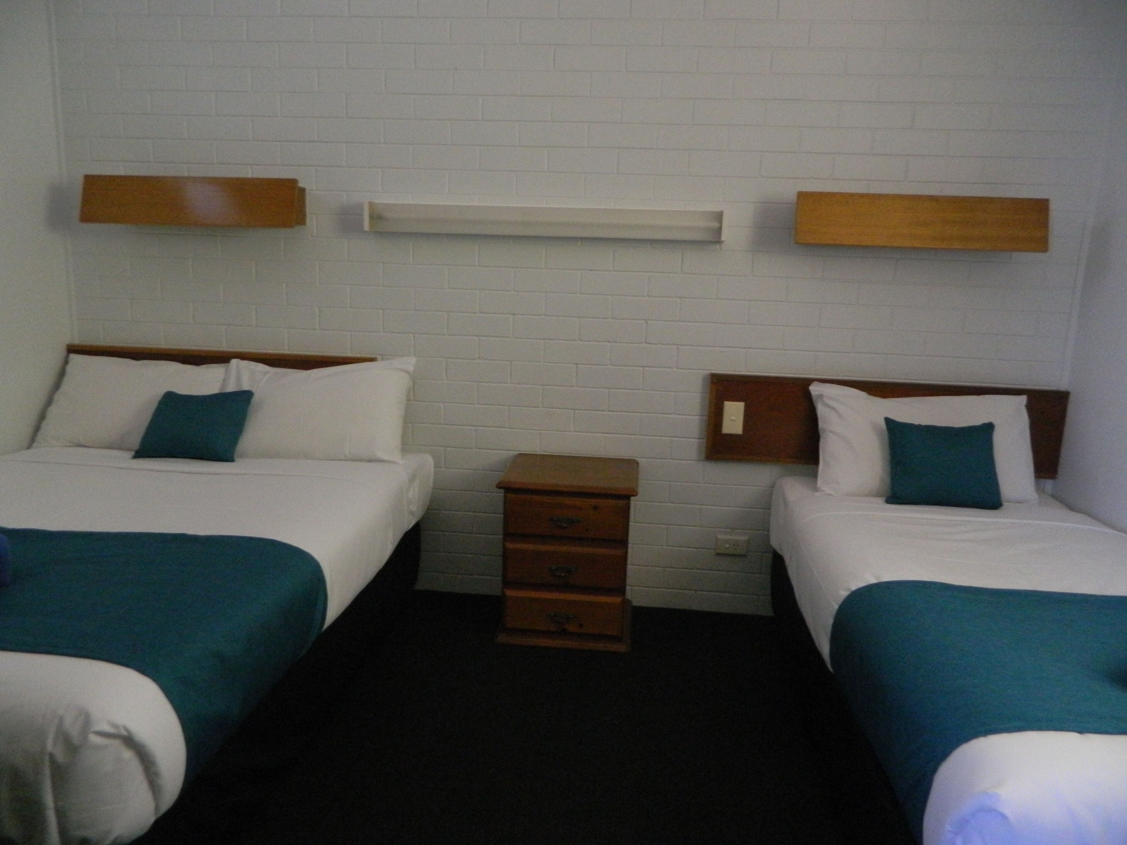 A hotel room with two beds, each with white bedding and teal pillows. There is a wooden nightstand with drawers in the center.