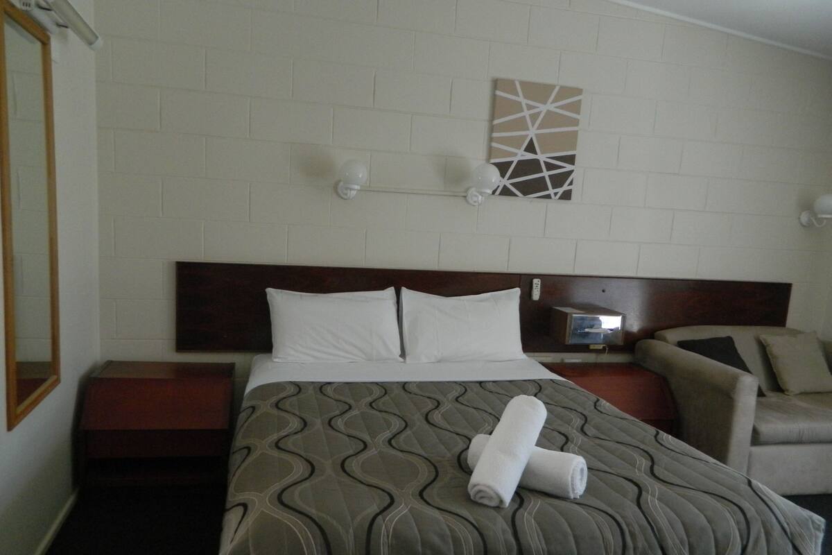 Una habitación de hotel con una cama, una silla, una mesita de noche, una lámpara de pared y un cuadro.