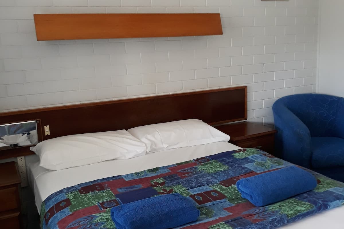 Una cama bien hecha con un colcha colorida, una silla azul y un cabecero de madera frente a una pared de ladrillo blanco.