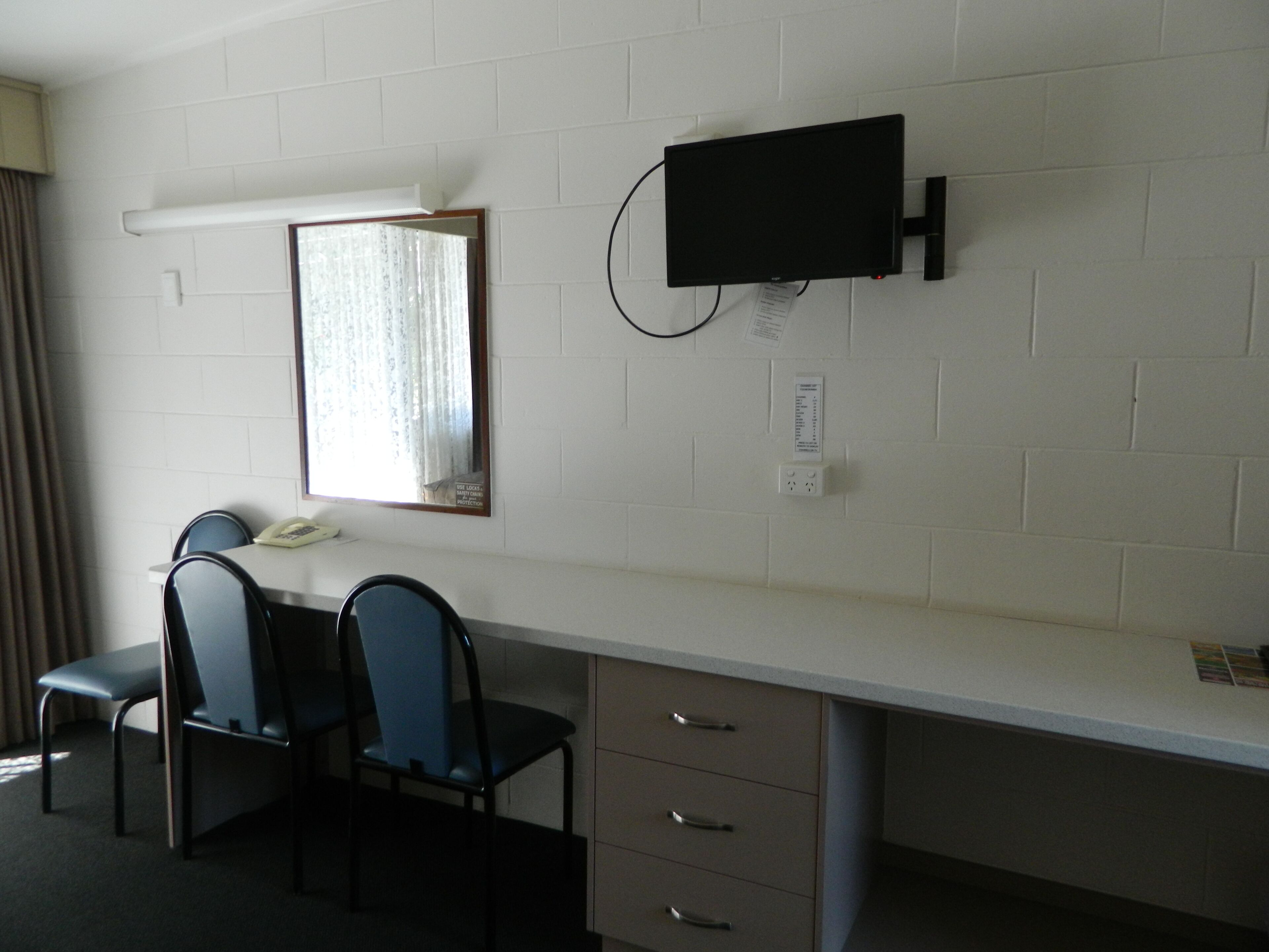 A hotel room with a desk, two chairs, a telephone, a television mounted on the wall, and a window with curtains.